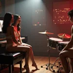 Two women in lingerie sit by a piano while a man plays drums in a dimly lit room.