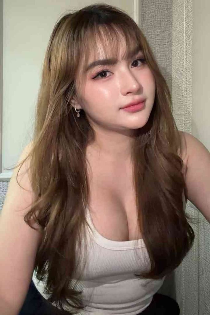 Young woman with long brown hair and bangs, wearing a white tank top.